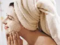 Kobieta z ręcznikiem na głowie nakłada na twarz peeling. To domowy sposób na gładką skórę.