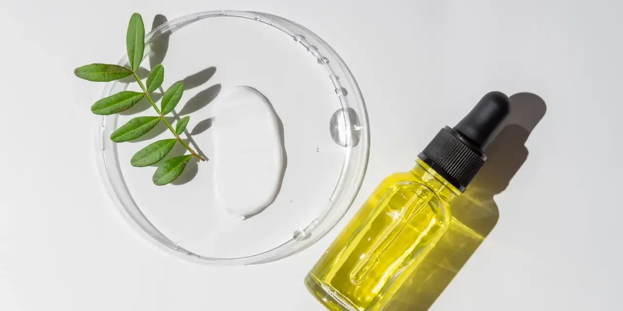 Kropla białego kremu i żółte serum w buteleczce z pipetką, obok gałązka z liśćmi. Składniki do walki z trądzikiem, w tym retinol.