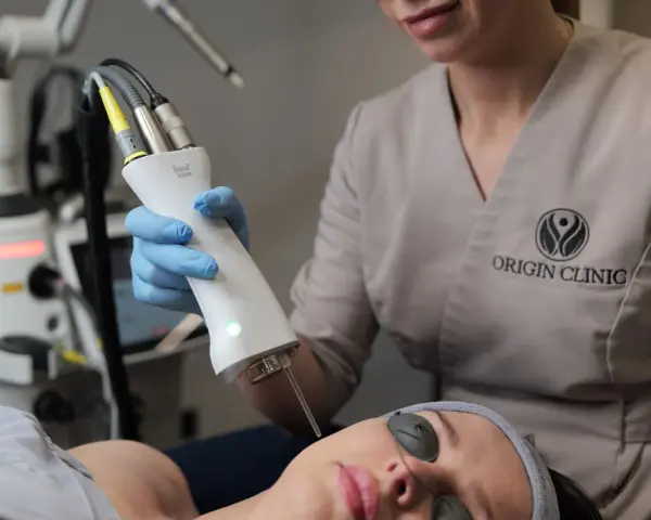 Zabieg laserem Fotona R35NX w Origin Clinic. Widoczna głowica lasera i pacjentka w okularach ochronnych. Oczekiwane spektakularne efekty odmładzające.