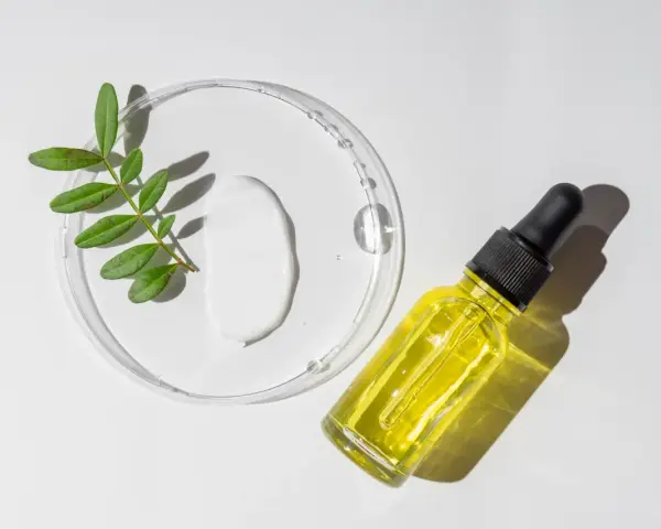 Kropla białego kremu i żółte serum w buteleczce z pipetką, obok gałązka z liśćmi. Składniki do walki z trądzikiem, w tym retinol.