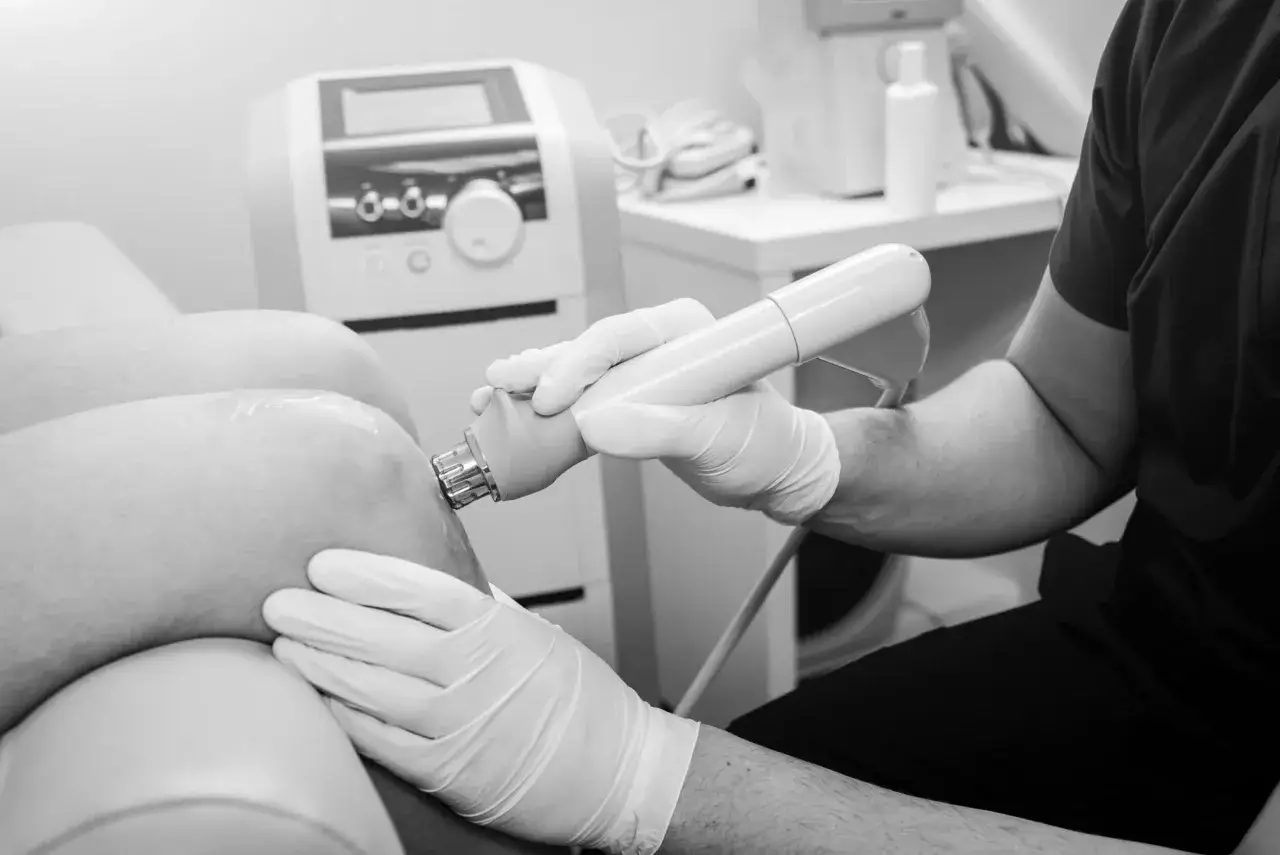 Fizjoterapeuta w rękawiczkach wykonuje zabieg fizjoterapii laserem na kolanie pacjenta. Czas zabiegu jest kluczowy dla skuteczności terapii.
