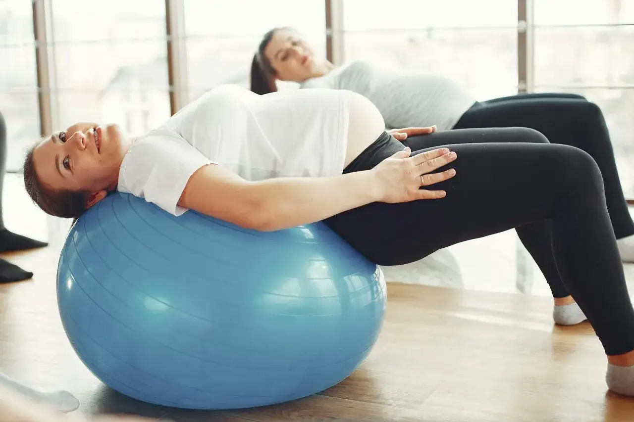 Kobiety w ciąży ćwiczą na piłce. Jedna z nich, z widocznym brzuchem po ciąży, opiera się o niebieską piłkę.